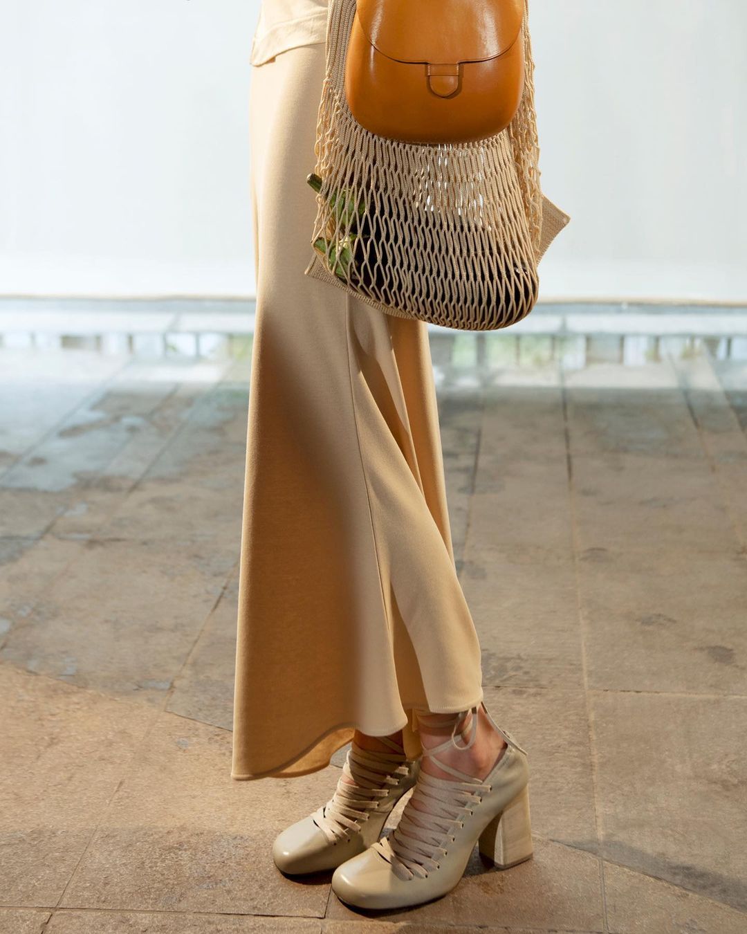 Beige heeled shoes, matching skirt, and tan purse in a net bag create a muted, fashionable look against a neutral background.