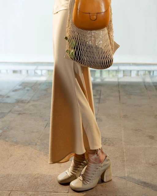 Beige heeled shoes, matching skirt, and tan purse in a net bag create a muted, fashionable look against a neutral background.