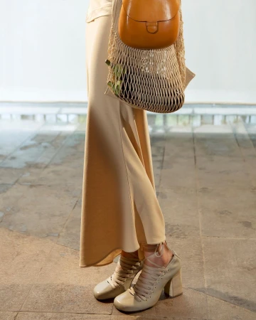 Beige heeled shoes, matching skirt, and tan purse in a net bag create a muted, fashionable look against a neutral background.