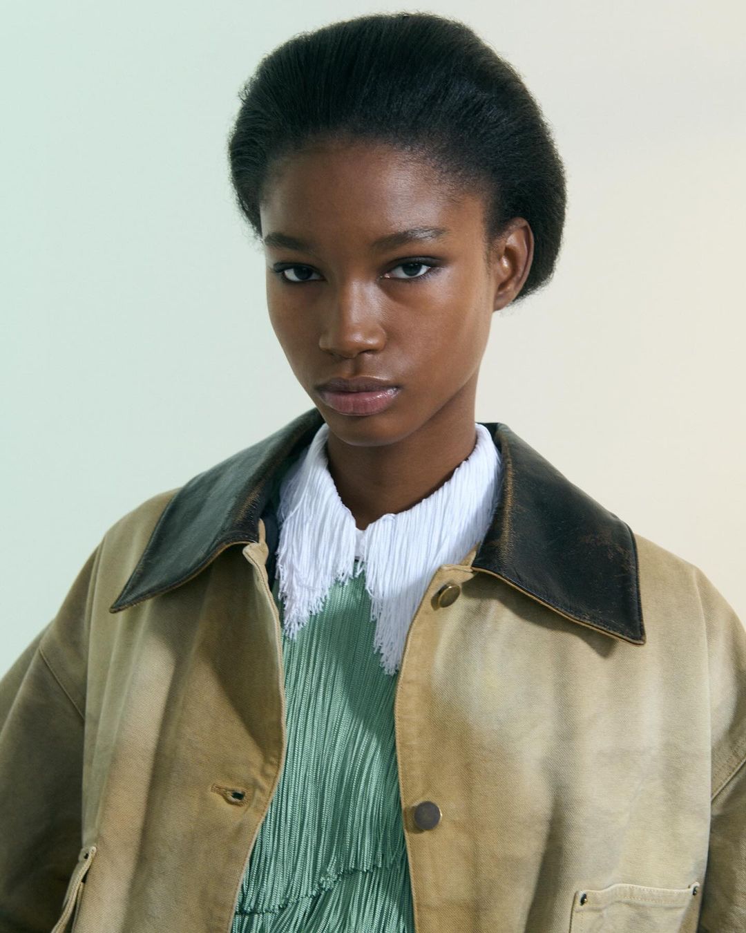 Portrait of a Black woman wearing a tan workwear jacket over a white collared shirt and a green top, standing indoors.