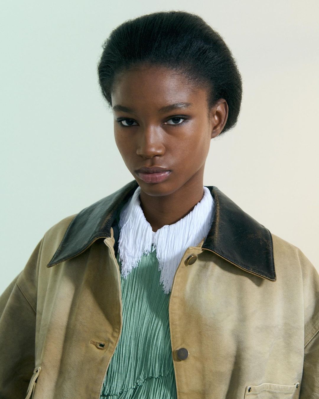 Portrait of a young Black woman in a tan jacket, wearing a green pleated dress and a white textured collar, posing in a photo studio.