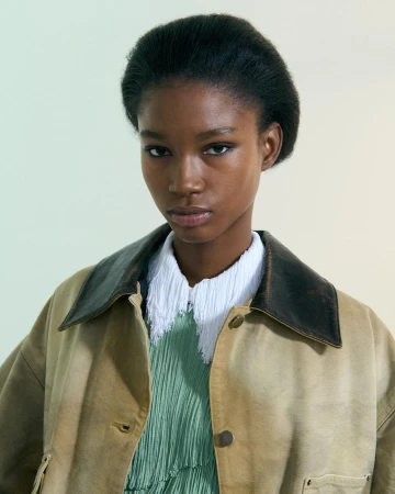 Portrait of a young Black woman in a tan jacket, wearing a green pleated dress and a white textured collar, posing in a photo studio.