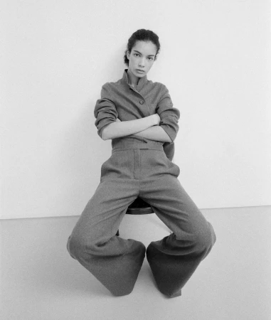 Model with arms crossed wearing a grey buttoned jacket and trousers seated on a stool against a plain background