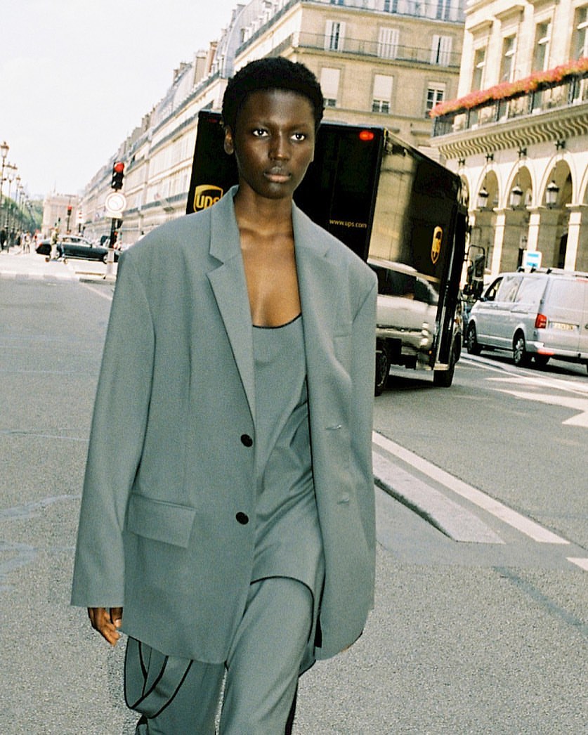 A woman in a grey suit walks down a city street with a UPS delivery truck in the background on a sunny day in an editorial fashion shot.