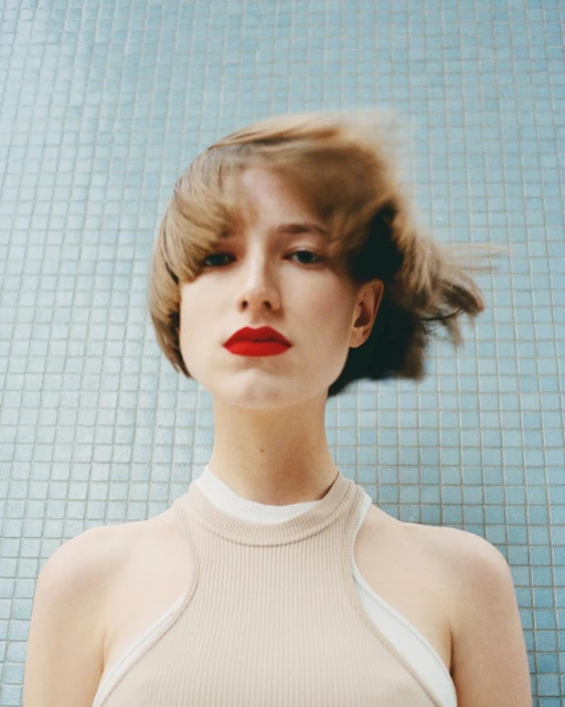 Close-up portrait of an adult female with red lipstick, styled short hair blowing in the wind, wearing a light tan top, in front of a tile wall.