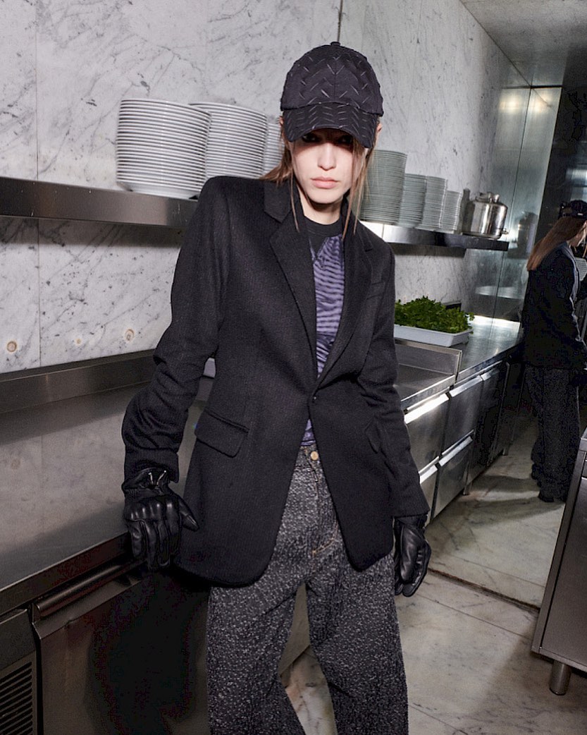 A woman in a black blazer and cap stands in a commercial kitchen, featuring stainless steel surfaces and neatly stacked white plates in background.