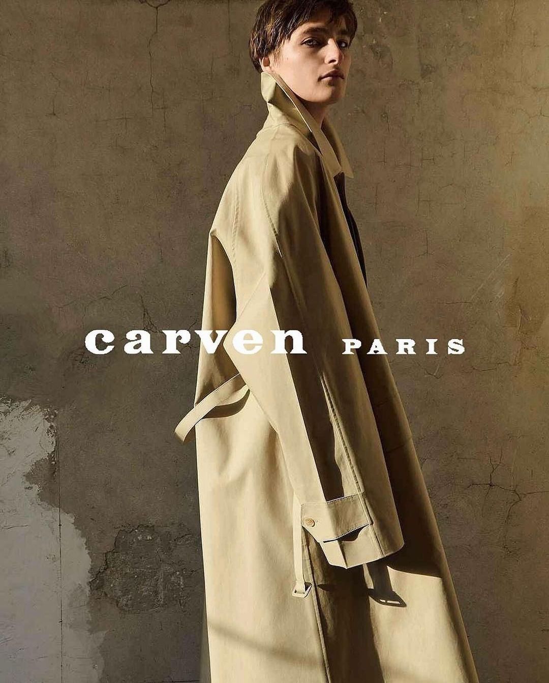 Adult woman wearing a tan overcoat, looking at the camera in a fashion setting. The photograph displays muted neutral colors.
