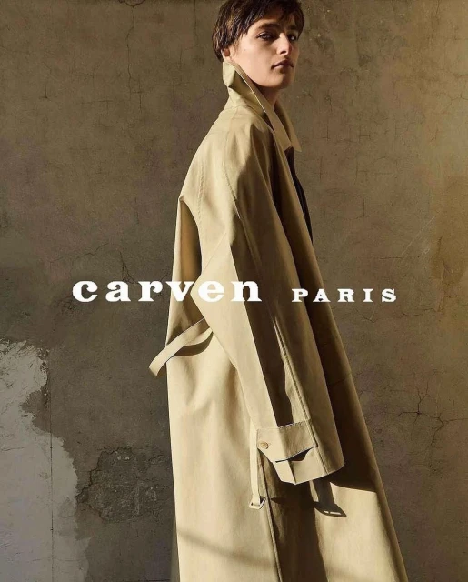 Adult woman wearing a tan overcoat, looking at the camera in a fashion setting. The photograph displays muted neutral colors.