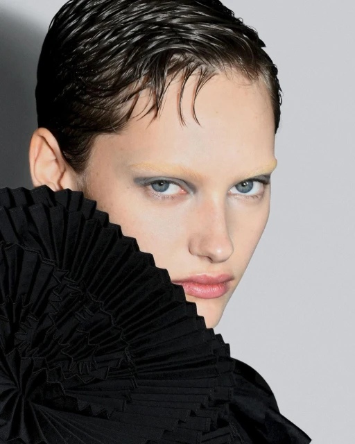 Close-up studio portrait of a light-skinned young adult female with short, slicked-back wet-look hair wearing black ruffled garment.