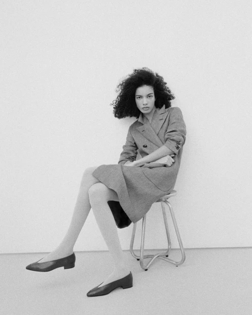 Black and white studio shot of a curly haired woman sitting on a stool, wearing a dress coat and shoes, with tights.