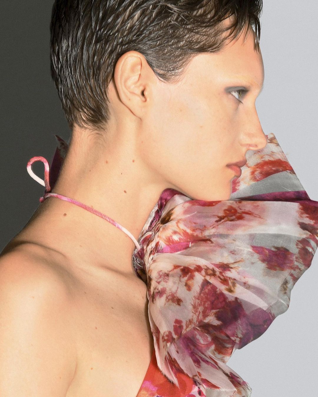 Profile shot of a fair-skinned adult female model with a short dark haircut, wearing a floral-patterned garment against a gradient gray background.