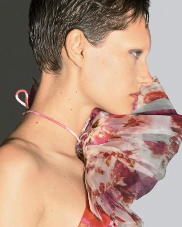 Profile shot of a fair-skinned adult female model with a short dark haircut, wearing a floral-patterned garment against a gradient gray background.