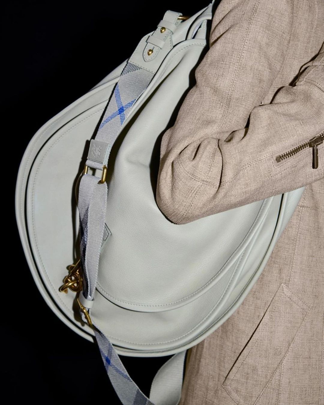 Close-up of a light blue leather handbag with a patterned strap, carried by a person in a beige coat against a dark backdrop.