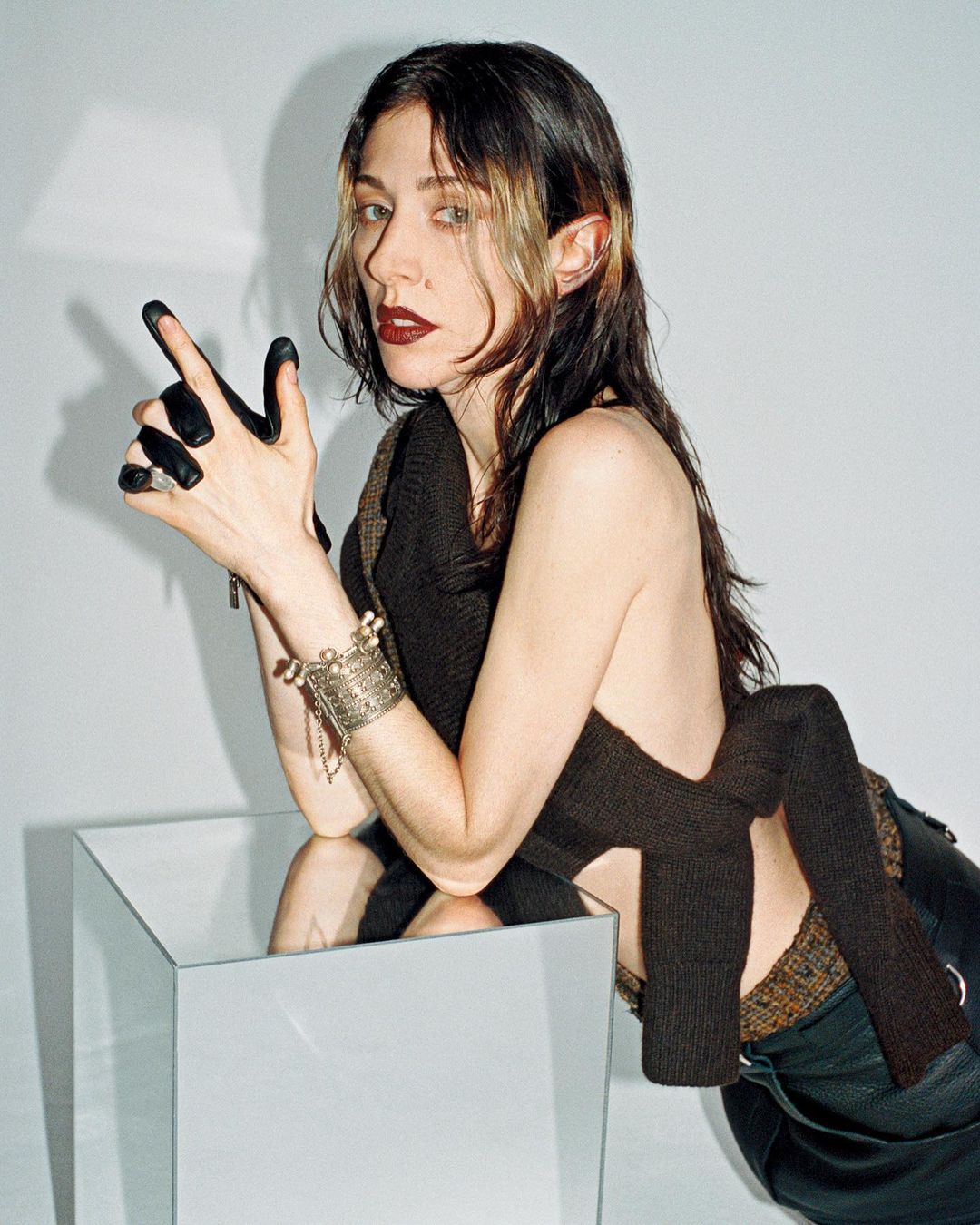 Portrait of a woman with dark hair and dark lipstick posing next to a reflective surface against a white backdrop in a studio.