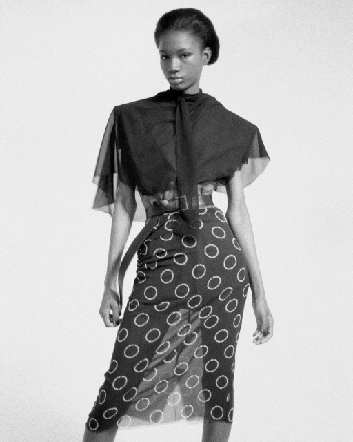Monochrome fashion shot of a Black female model wearing a black and white polka dot skirt and a sheer black shawl, posing in a studio.