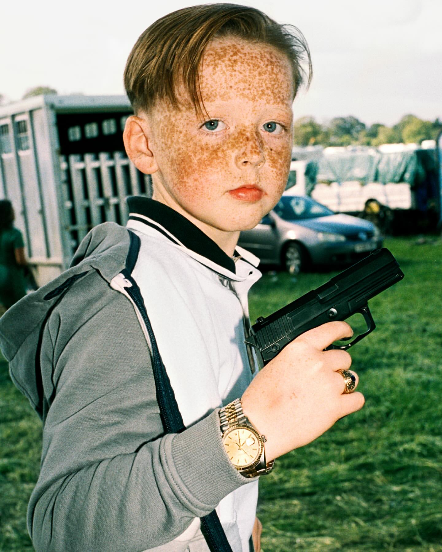 A young boy with freckles holds a black handgun while wearing a gold watch and rings, with trailers in the background and a green lawn.