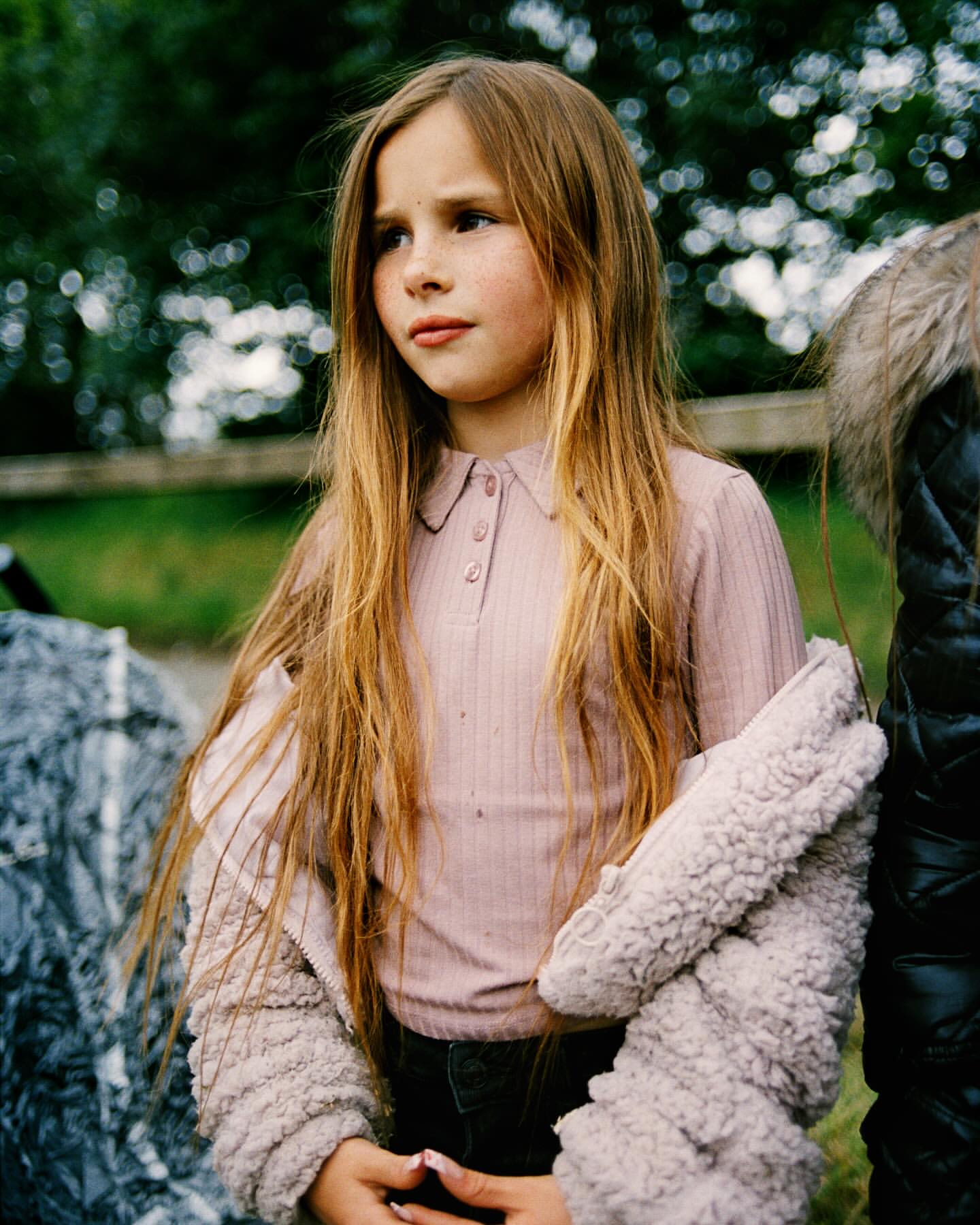 Portrait of a young girl with long brown hair wearing a pink long-sleeved shirt and beige coat, posing outdoors with a blurred background.