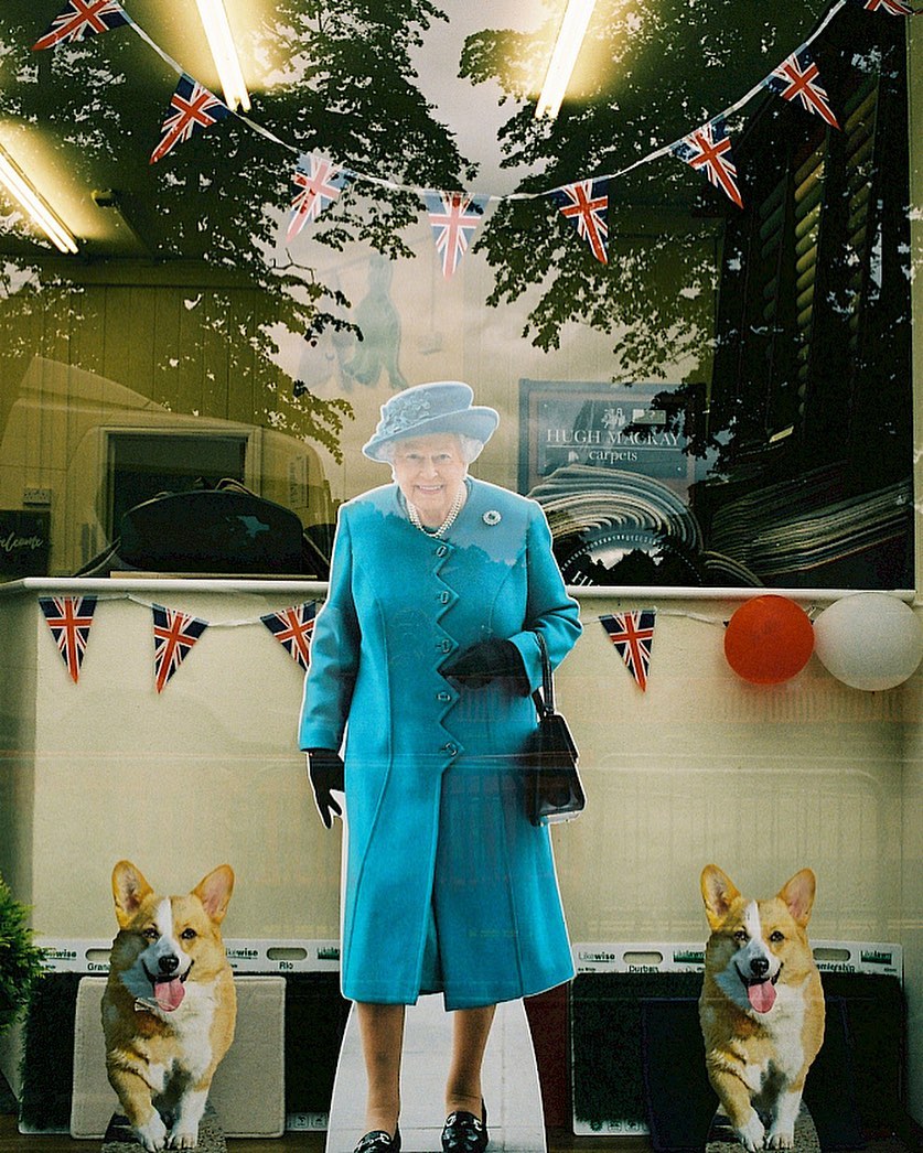Queen Elizabeth II cardboard cutout stands with two corgi cutouts under Union Jack bunting with tree shadows in the background.