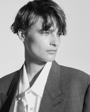 Close-up monochrome portrait of a model with short dark hair wearing a blazer and button-up shirt against a plain background.