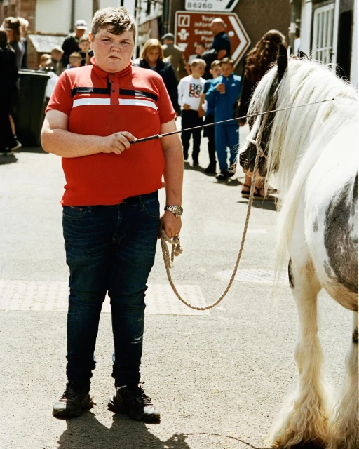 Young man stands in the street next to a pony with a rope, wearing a red shirt and jeans, surrounded by a crowd of people.