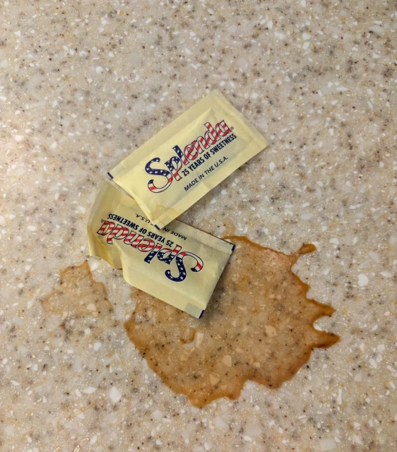 Two Splenda sweetener packets sit on a speckled countertop next to a spill of sweetener crystals.