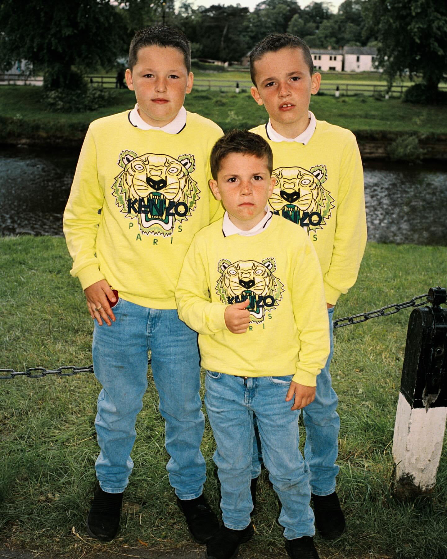 Three children wearing matching yellow sweatshirts and blue jeans pose outdoors in front of grass, a body of water and a building.