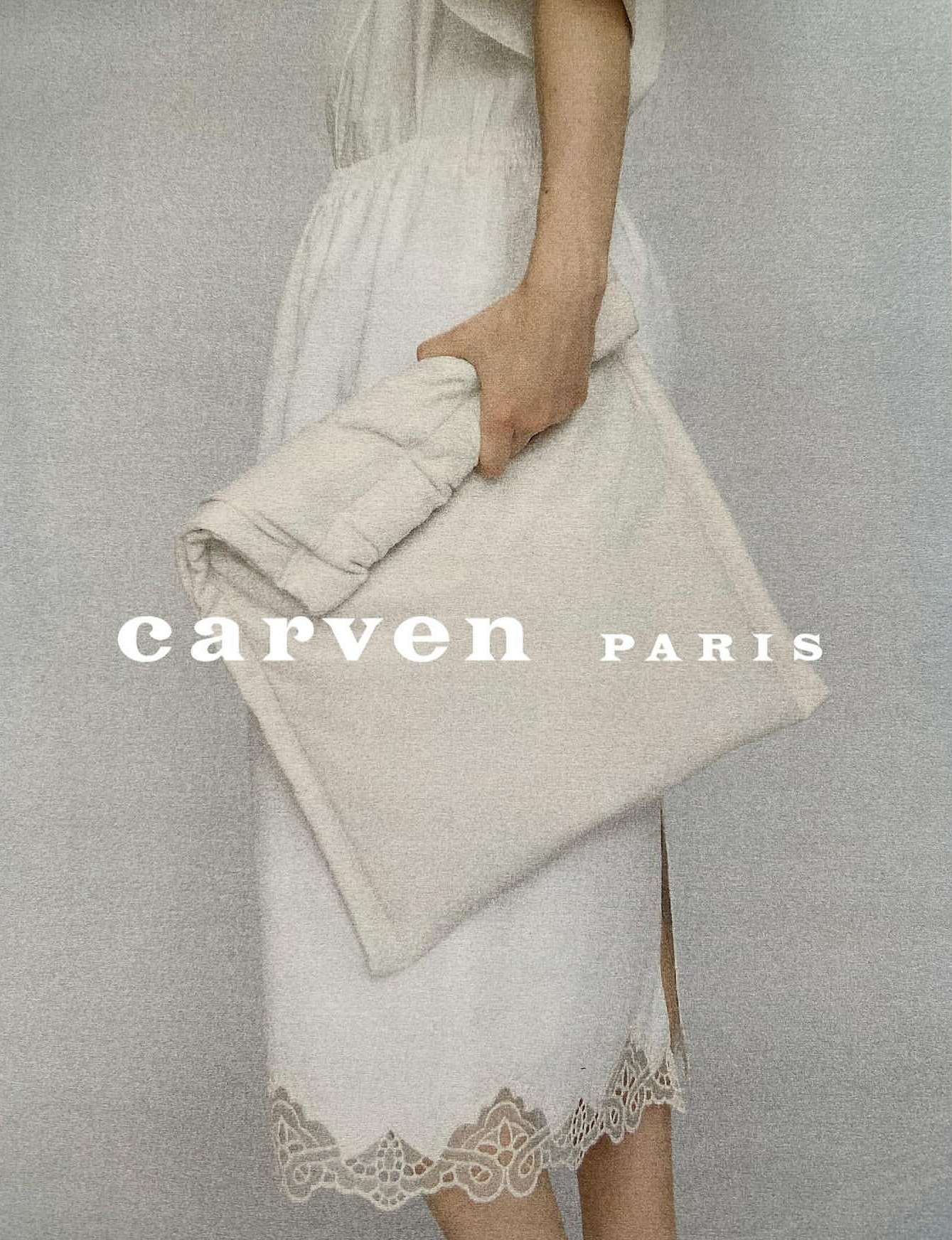 Woman in white dress holding a soft square Carven Paris clutch bag against a plain background in a fashion studio setting.