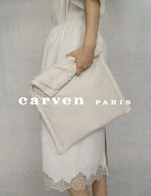 Woman in white dress holding a soft square Carven Paris clutch bag against a plain background in a fashion studio setting.