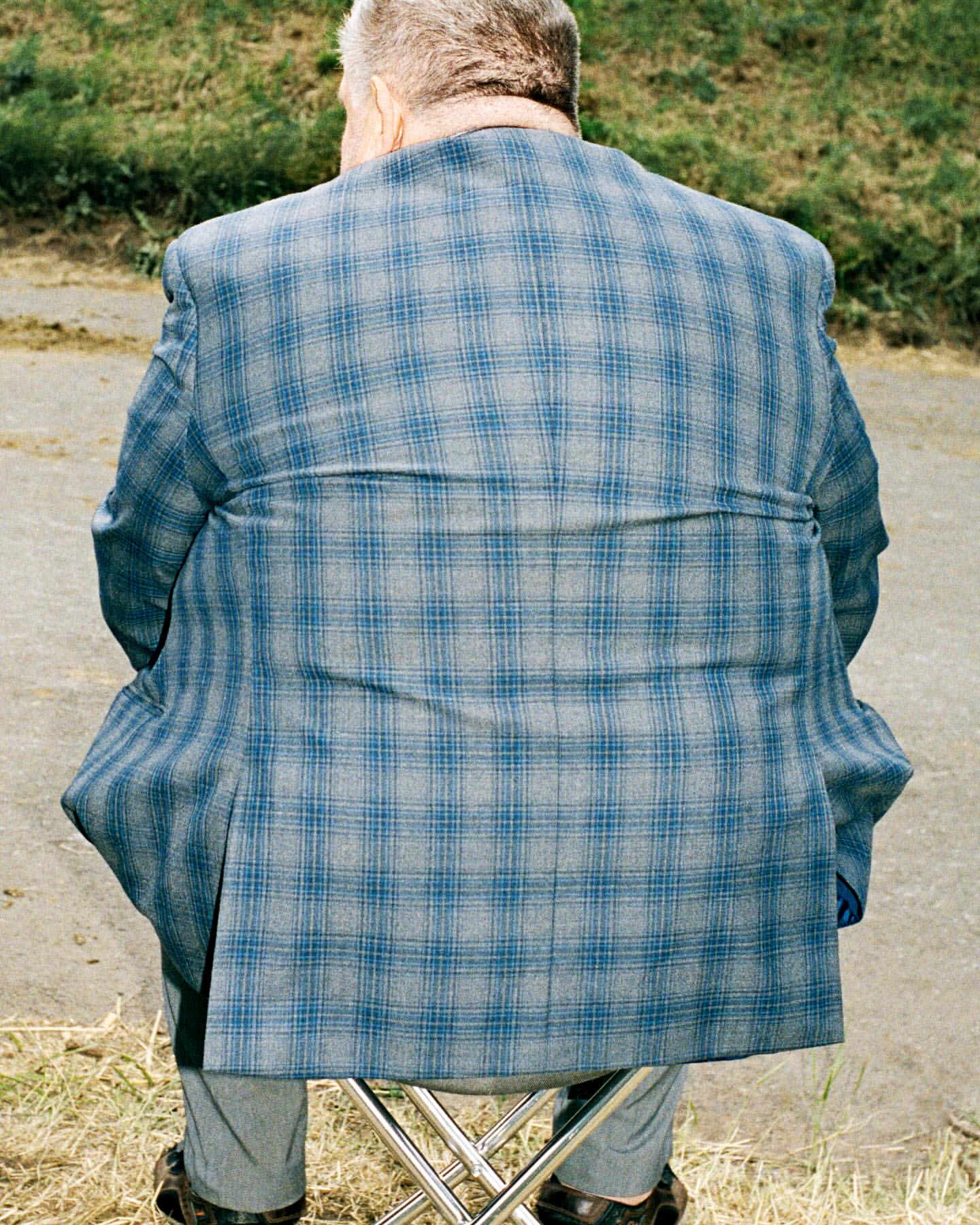 Elderly man in a blue plaid jacket sits on a folding stool outdoors, back to the viewer. Neutral and muted colors, natural lighting.