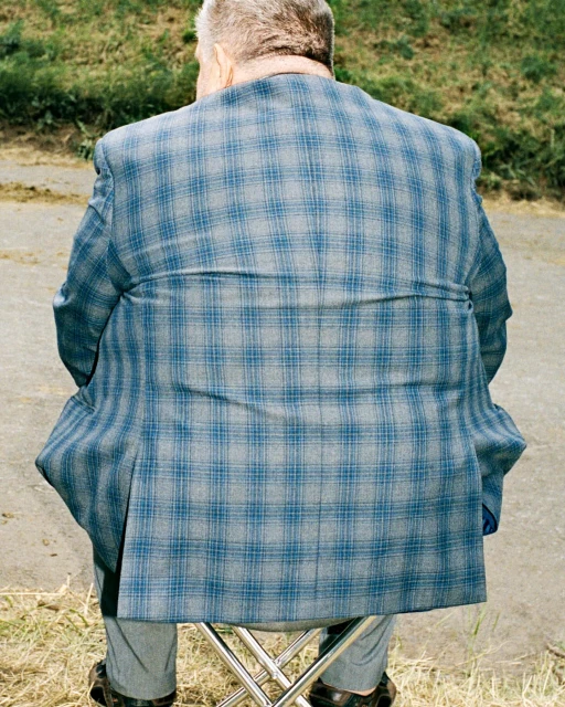 Elderly man in a blue plaid jacket sits on a folding stool outdoors, back to the viewer. Neutral and muted colors, natural lighting.