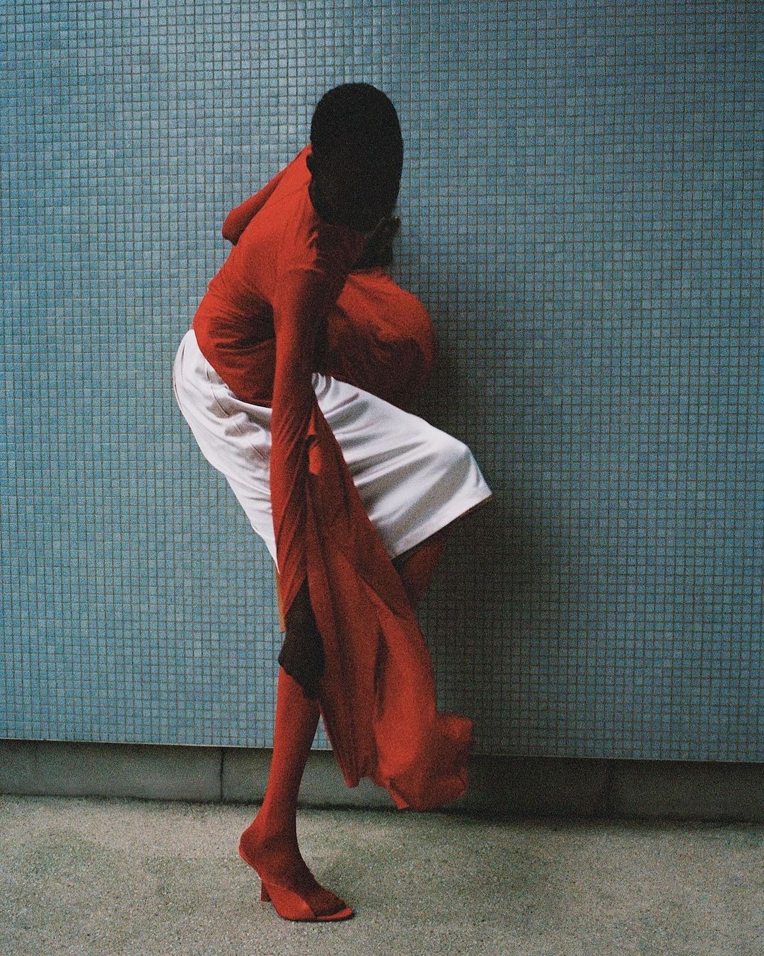 Person in red top and hite shorts posing against a blue tiled wall, wearing matching red socks and high-heeled shoes in a modern fashion shot.