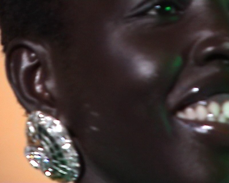 Close-up on an adult female with dark skin, wearing large earrings and a glossy lip. The background features green and orange tones.