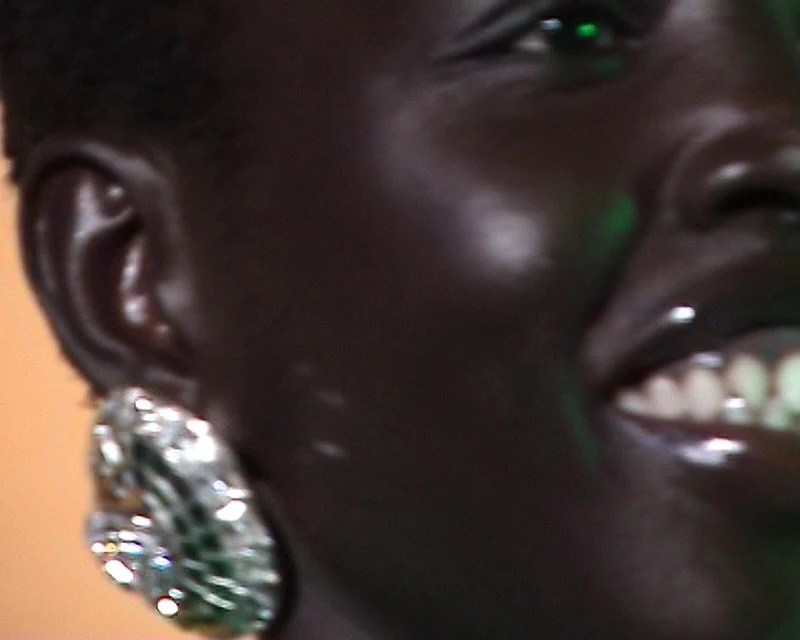 Close-up on an adult female with dark skin, wearing large earrings and a glossy lip. The background features green and orange tones.