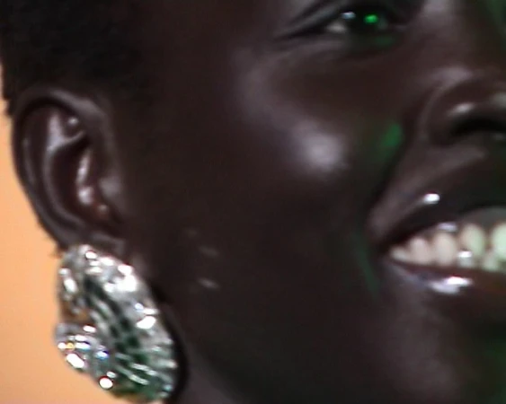 Close-up on an adult female with dark skin, wearing large earrings and a glossy lip. The background features green and orange tones.
