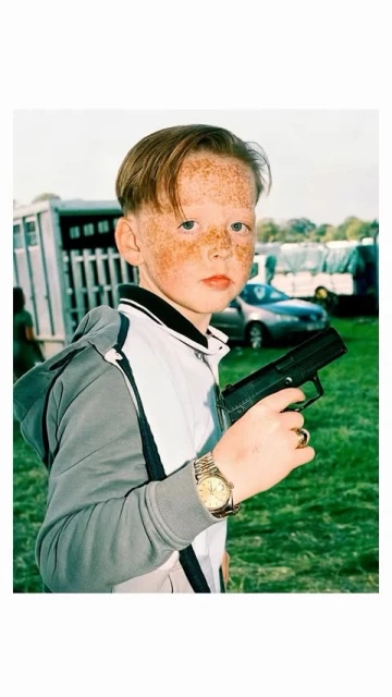 Young boy with freckles poses outdoors holding a black toy gun, wearing a grey and white tracksuit, gold watch and gold ring.