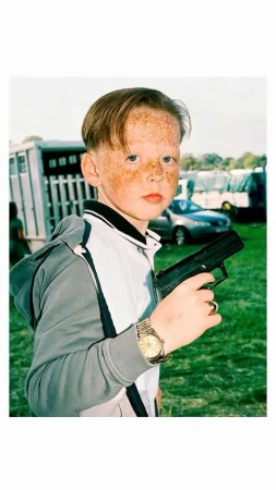 Young boy with freckles poses outdoors holding a black toy gun, wearing a grey and white tracksuit, gold watch and gold ring.