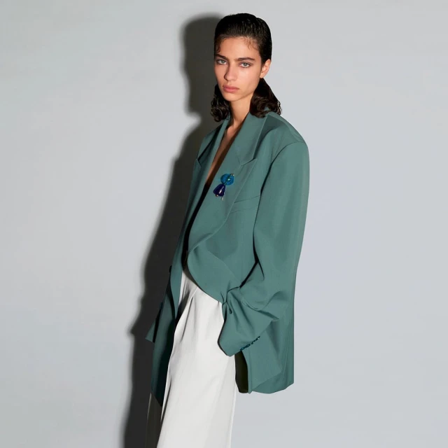 A woman poses indoors wearing a teal blazer and white pants, looking directly at the camera with a neutral expression.