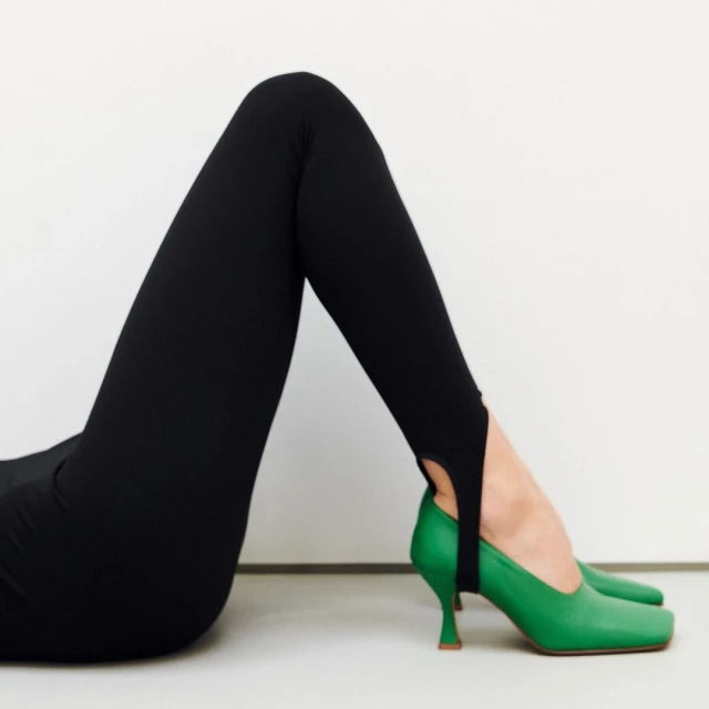 Woman in black leggings and green high heels lies against a white background in a fashion studio shot for clothing advertisement.