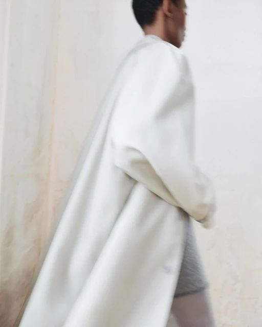 Fashion shot of an adult female model walking indoors wearing a light beige overcoat, grey dress and tights against a white wall.
