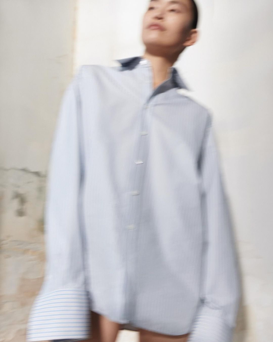 Blurry image of a slender Asian woman in an oversized blue-striped dress shirt posing in front of a textured, light-colored wall.