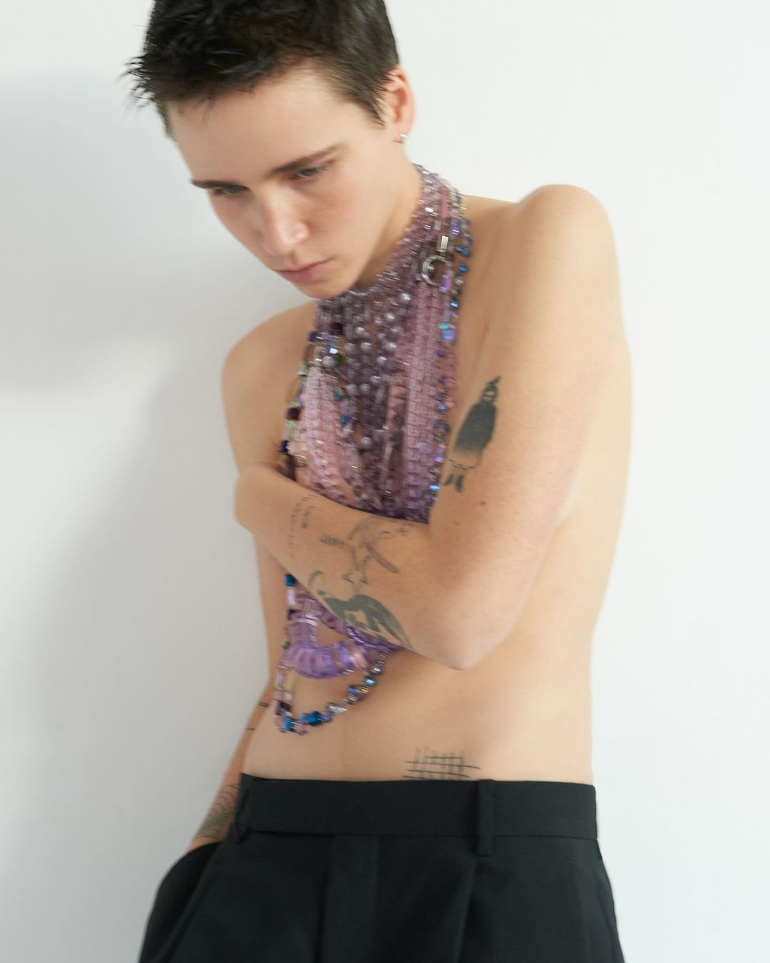 Nude-shouldered person wearing a decorative beaded covering around their neck and upper chest, paired with black trousers and tattoos.
