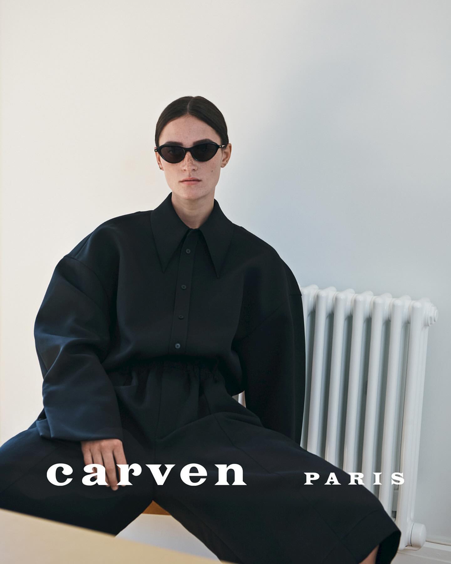 Model with dark sunglasses wearing an oversized black shirt and trousers against a plain wall with a white radiator.