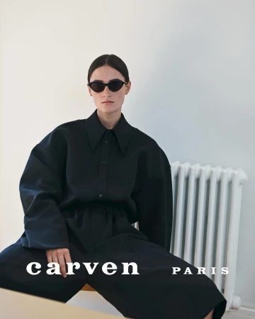 Model with dark sunglasses wearing an oversized black shirt and trousers against a plain wall with a white radiator.