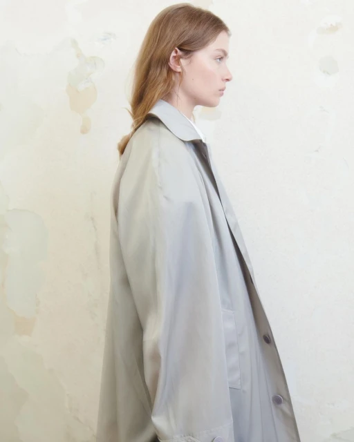 Profile portrait of a young fair-skinned female in a light grey trench coat against a simple off-white textured background.