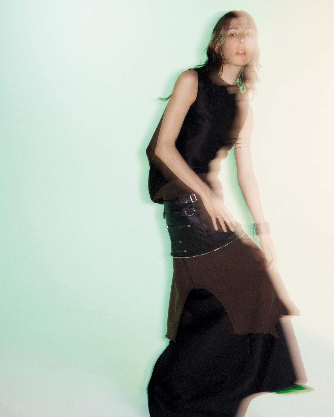 A woman in a sleeveless black top and layered skirt poses in front of a blurred green background, showcasing a unique fashion style.