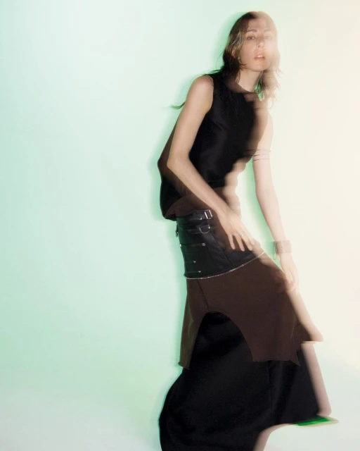A woman in a sleeveless black top and layered skirt poses in front of a blurred green background, showcasing a unique fashion style.
