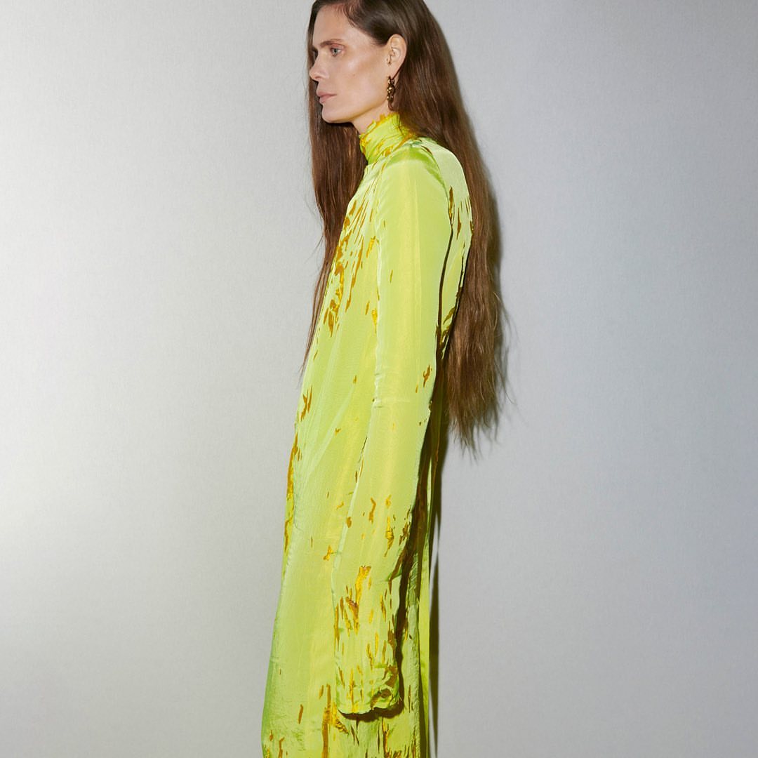 Side profile studio shot of an adult woman with long brown hair wearing a neon yellow, long-sleeved dress with gold accents.