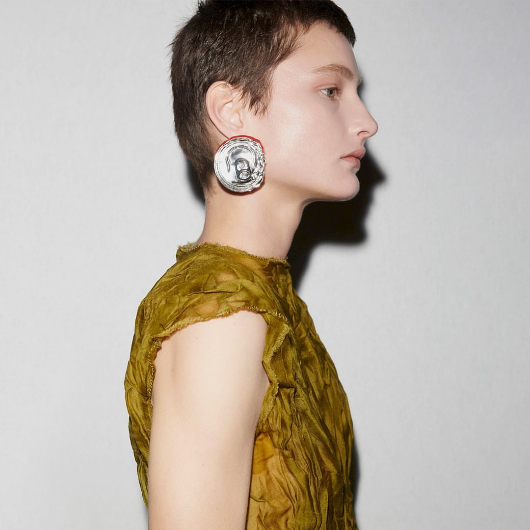 Woman with short hair wearing a brown top and large earrings posing in a side view in front of a neutral background. Fashion shot.