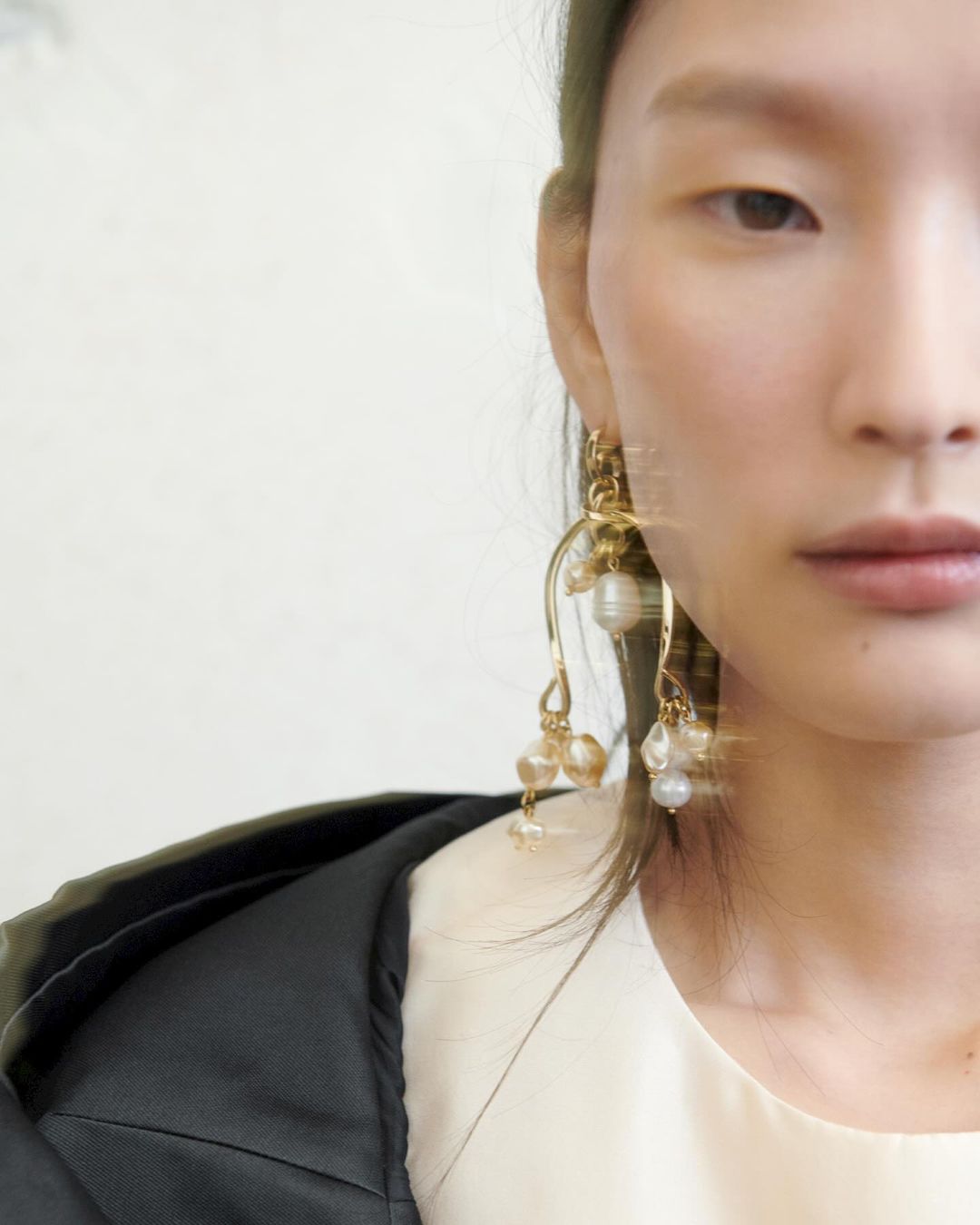 Close-up of an Asian female model with pearl drop earrings wearing a black blazer and cream-colored top in a studio setting.