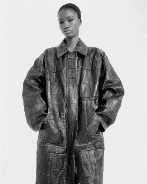 A woman in a black headscarf and a patchwork leather trench coat stands in a studio with her hands in her pockets.